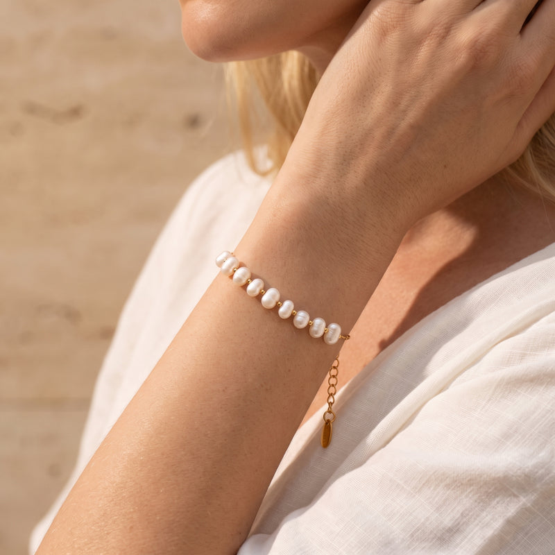 Gold-tone pearl station bracelet worn on a wrist over white fabric, with its extender chain hanging below.