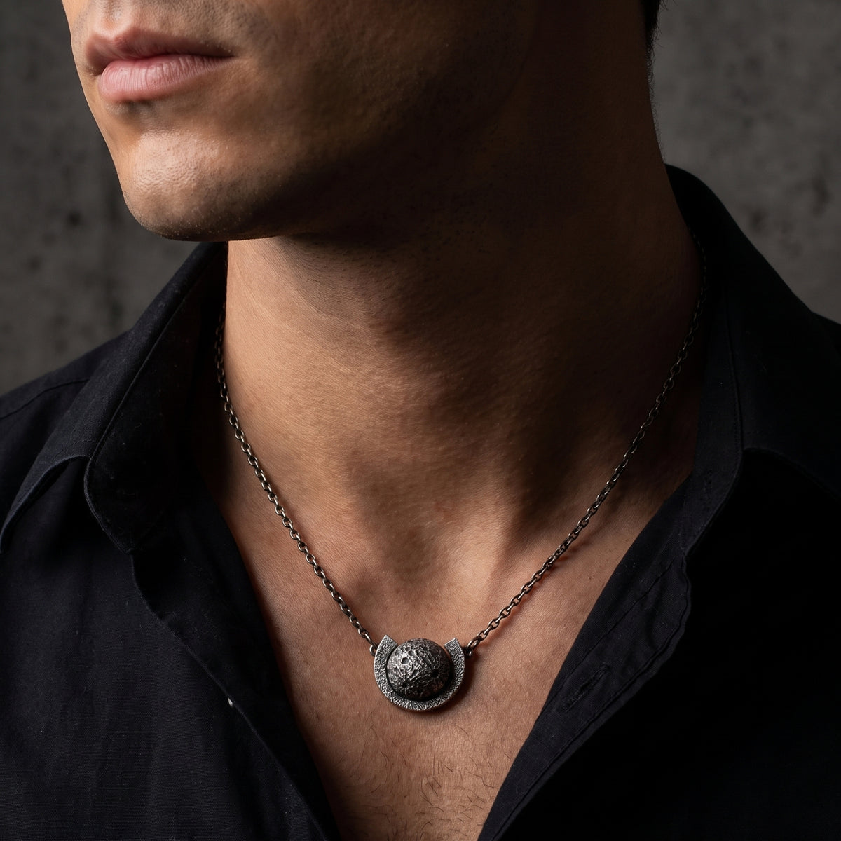 Close-up view of a man wearing the silver cratered moon necklace with an open black shirt collar.