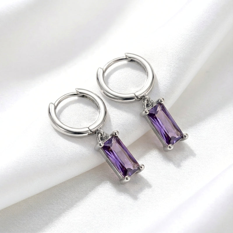 Sterling silver hoop earrings with emerald cut simulated amethyst drops resting on white fabric.