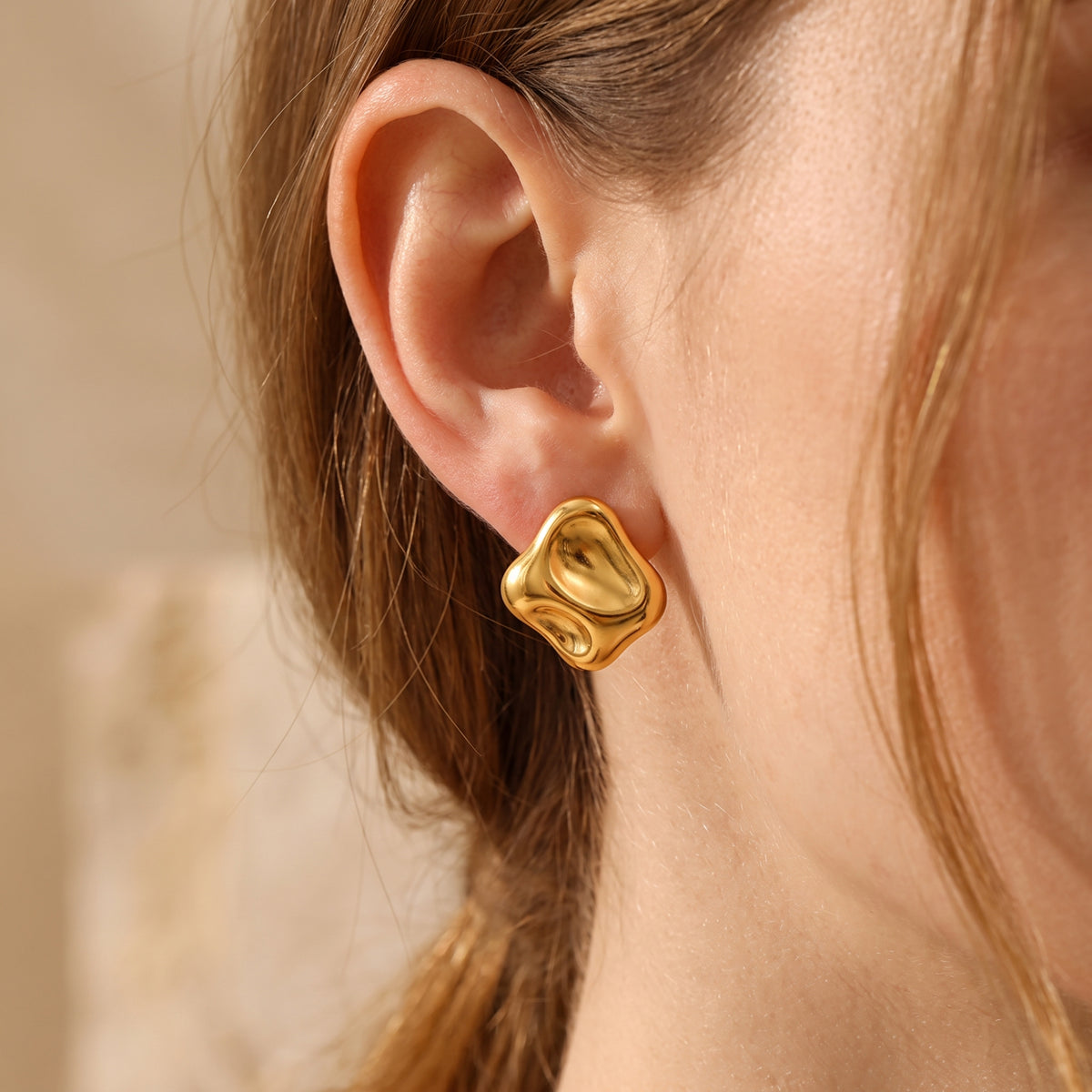 Close-up of one polished gold-tone sculptural square stud earring worn on an ear.