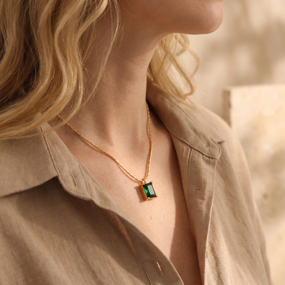 Model wearing a gold-tone necklace with an emerald-green rectangular pendant over a beige button-up shirt.