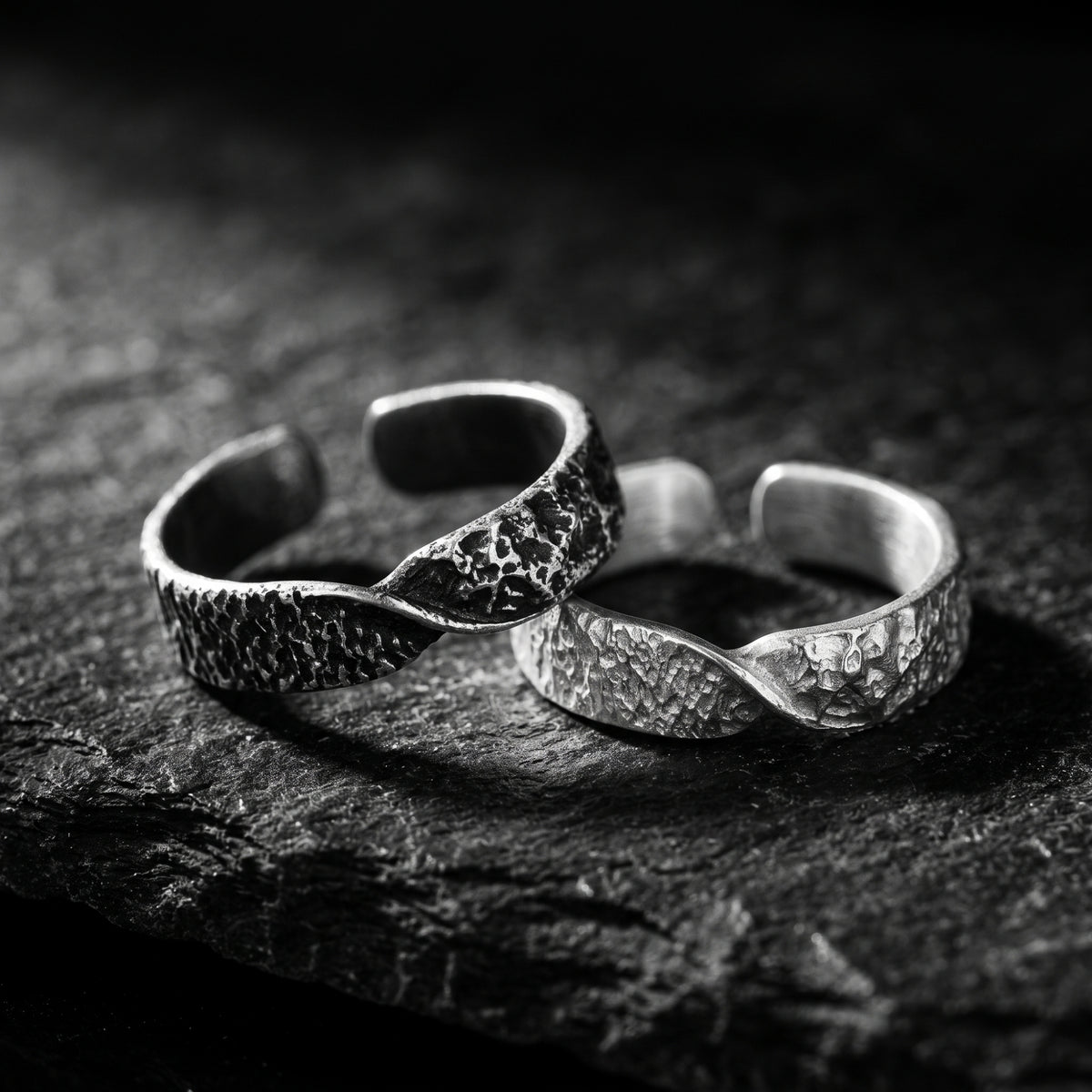 Dark oxidized and light silver hammered twist rings resting together on a textured black stone surface.