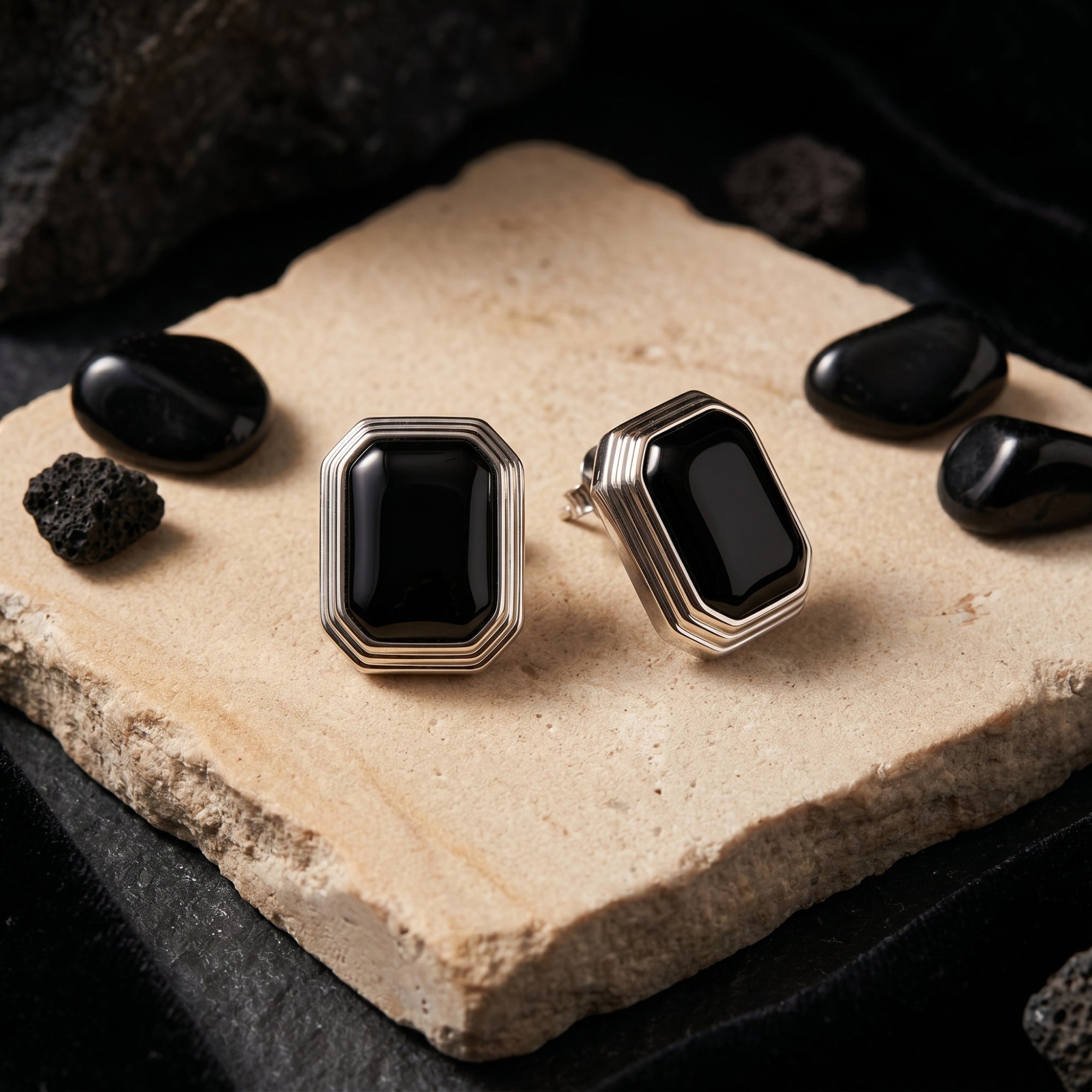 Two square cut natural black onyx earrings in silver, resting on a textured stone surface with black decorative rocks.