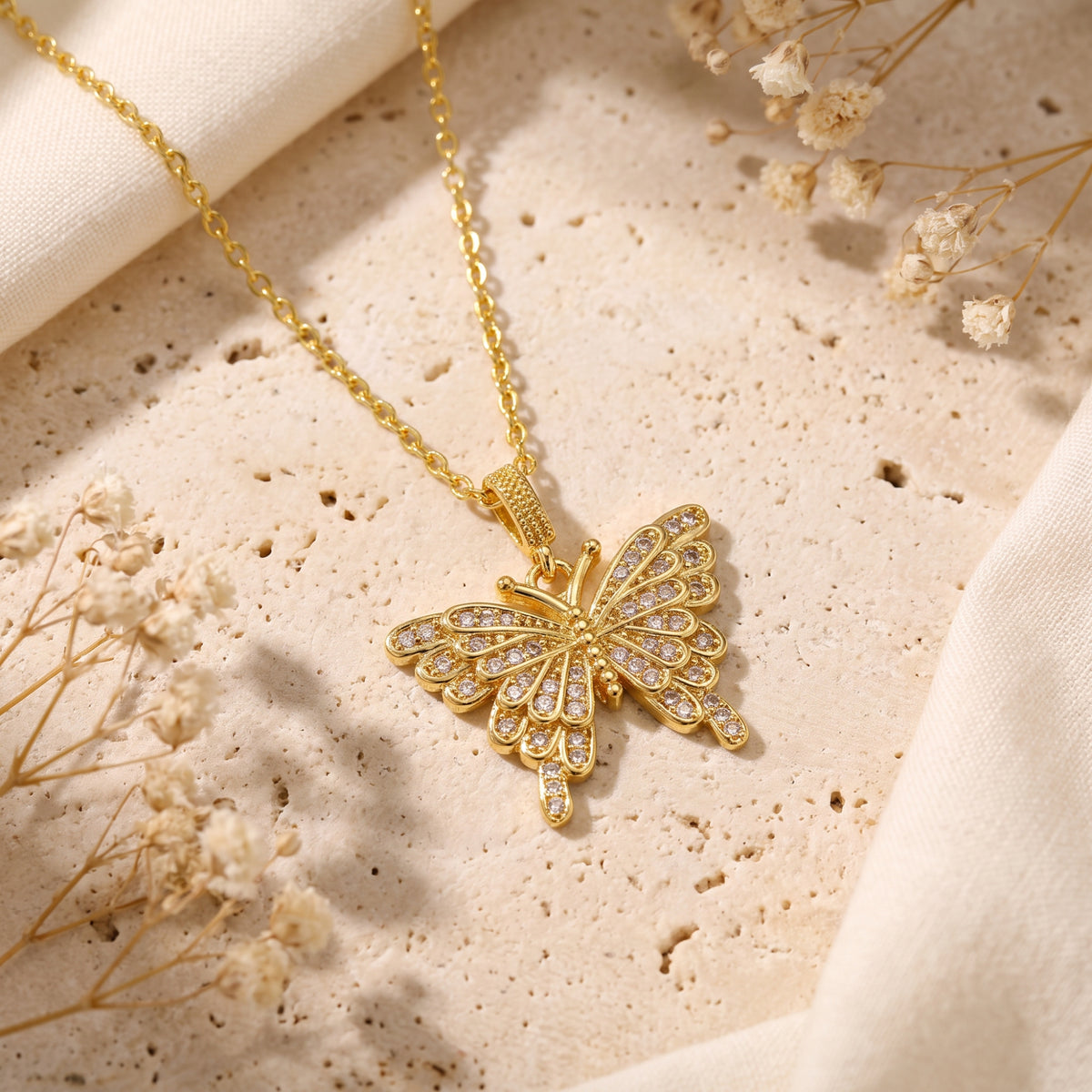 Gold-tone butterfly pendant with clear stones resting flat on a beige porous stone surface beside dried flowers.