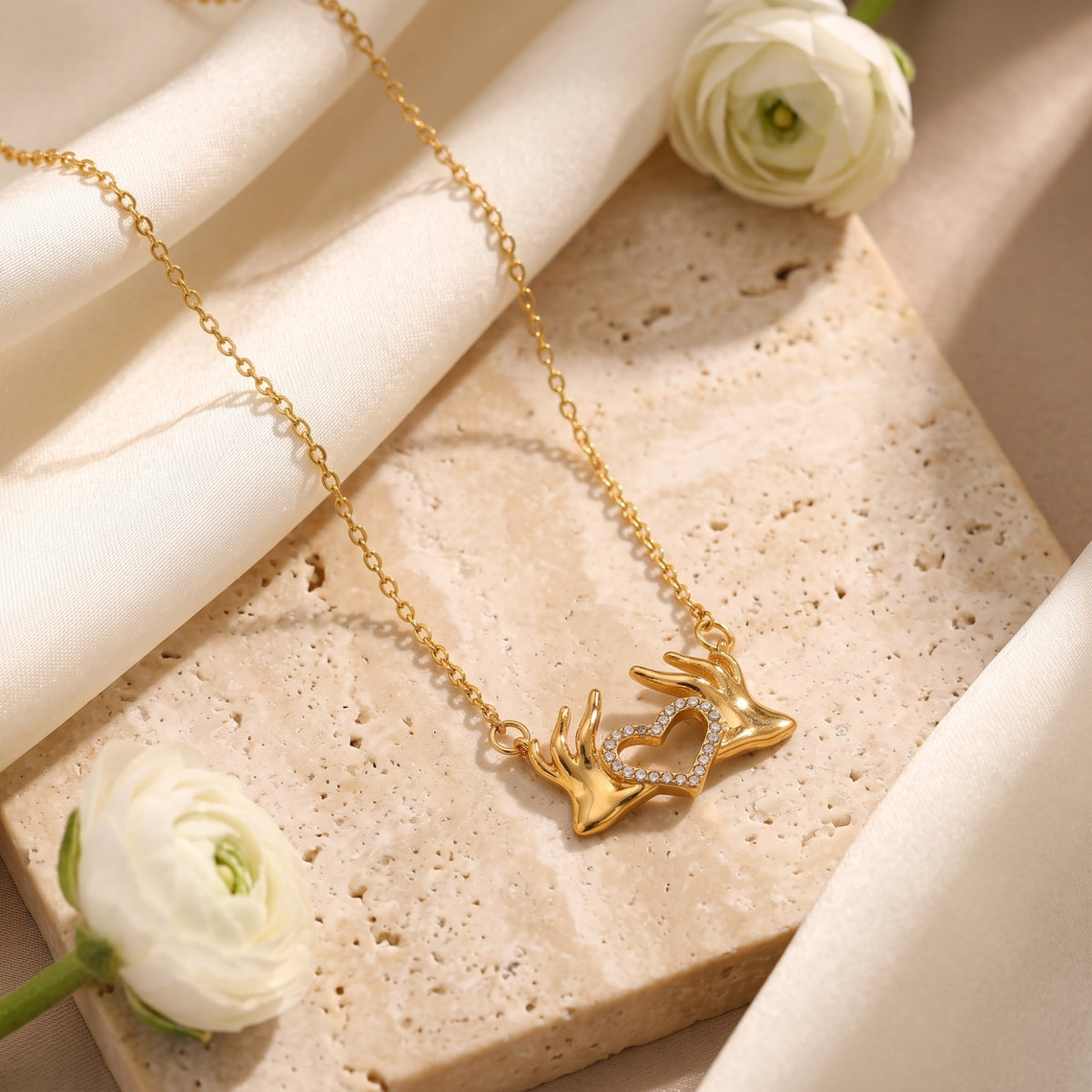 Gold-tone heart hands pendant necklace arranged diagonally on travertine with white flowers and draped silk.