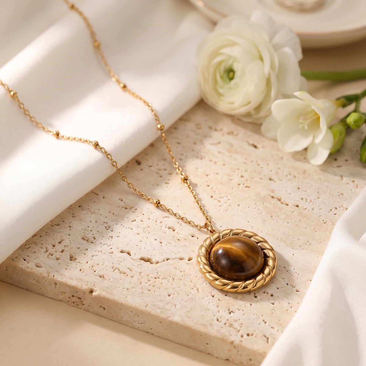 Gold-tone station chain with a round brown cabochon pendant laid on a porous beige slab beside white flowers