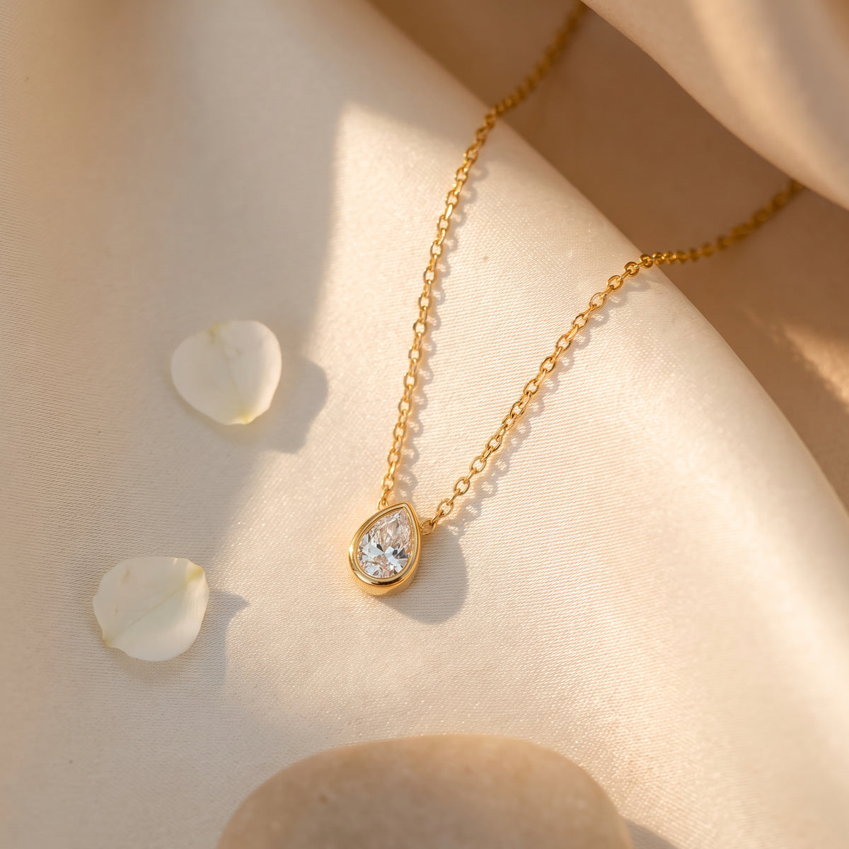 Gold-tone pear-cut pendant necklace arranged diagonally on cream fabric beside white petals.