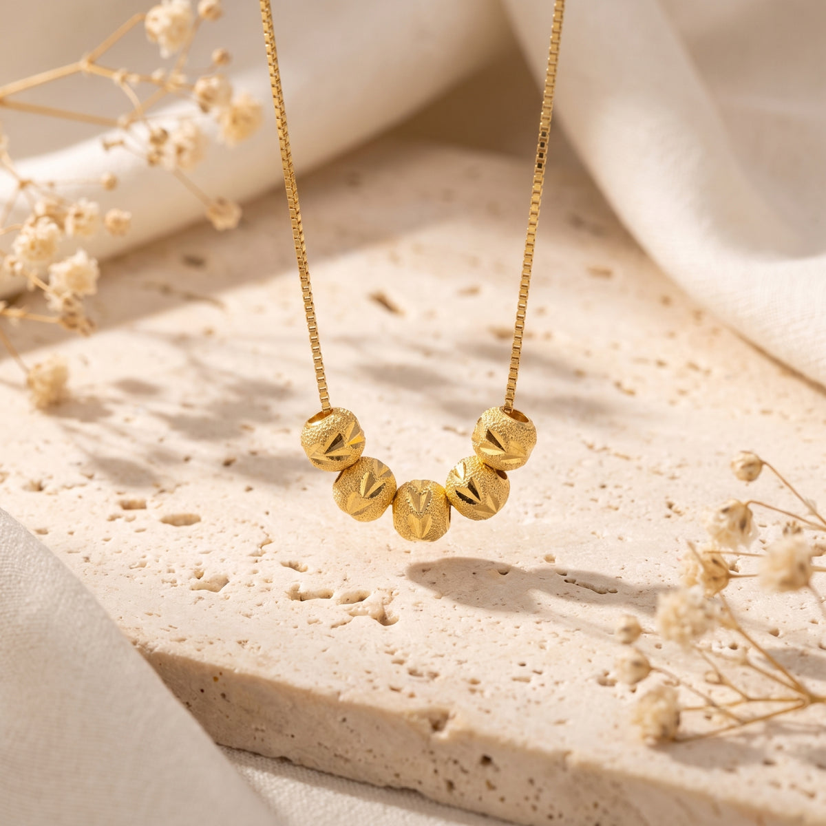 Close-up of five textured gold-tone beads suspended from a box chain above a stone slab.