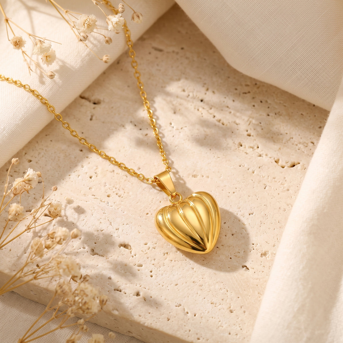 Angled view of a gold-tone ribbed heart pendant resting on textured stone beside delicate white florals.