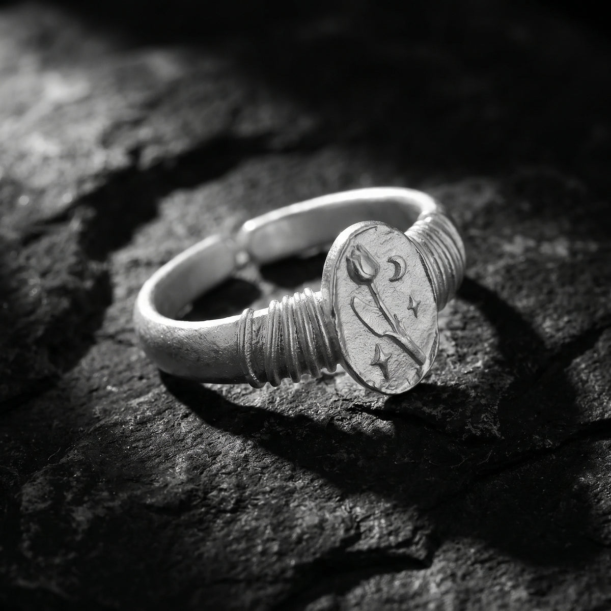 Angled silver tulip signet ring on dark stone, with its open adjustable band visible behind the engraved face.