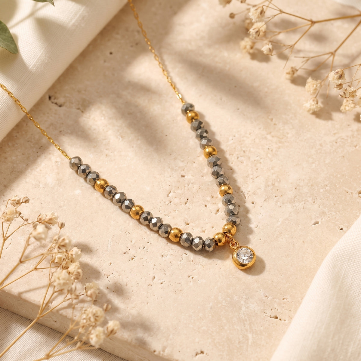 Overhead view of a gold-tone necklace with dark metallic beads and a round bezel-set pendant on a light stone surface.