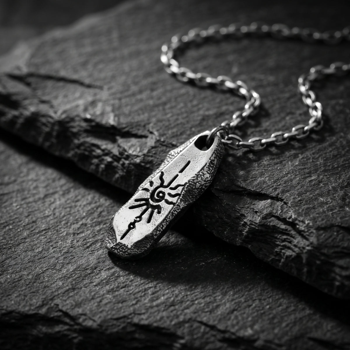 Angled silver pendant with an embossed solar motif resting on a dark slate surface beside its chain.