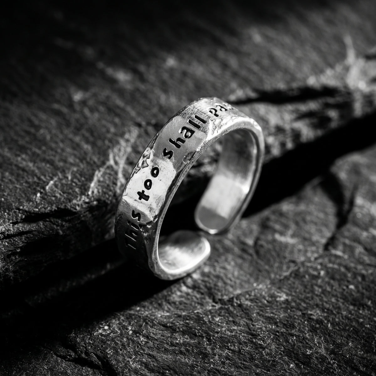 Angled view of the textured silver open-band ring on dark stone, showing the gap and engraved phrase.