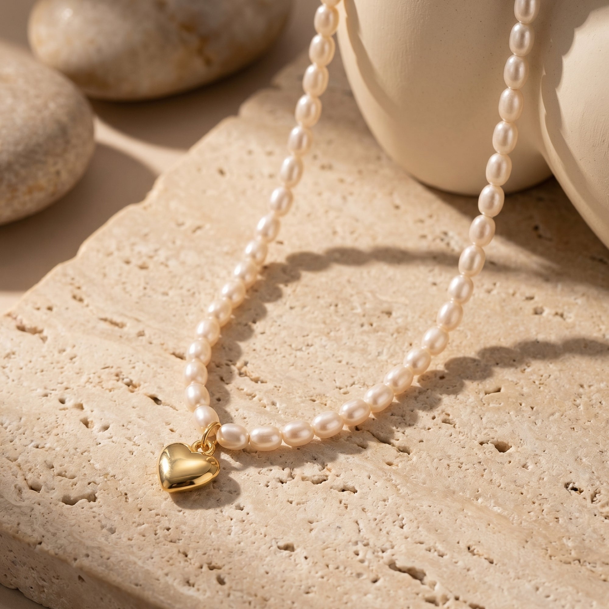 18K gold plated heart charm and white oval synthetic pearl necklace styled on a neutral stone background.