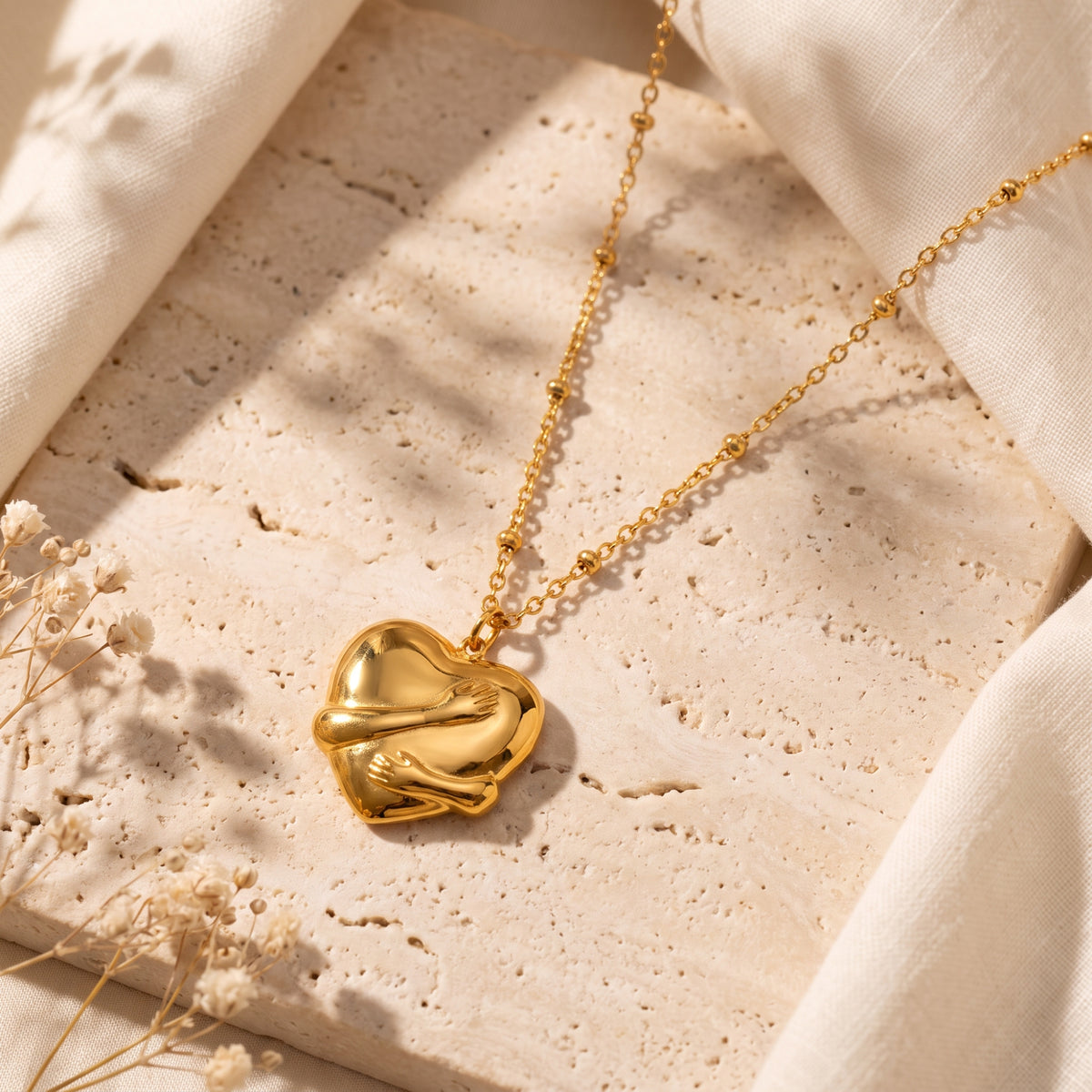 High-angle view of the gold-toned heart pendant with sculpted hands, resting on a beige travertine slab.