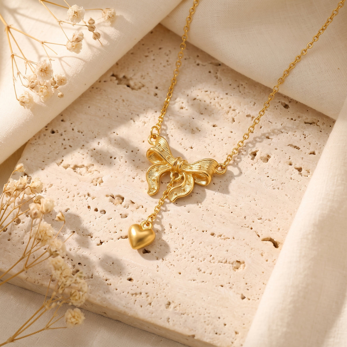 Gold-tone bow and heart pendant necklace displayed on a textured stone surface with fabric and dried flowers.