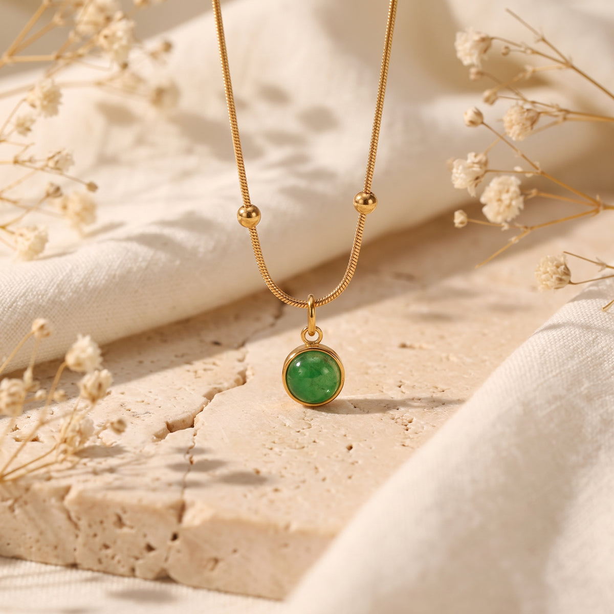 Close-up of a gold-tone snake chain with sphere stations and green cabochon pendant on a porous stone surface.