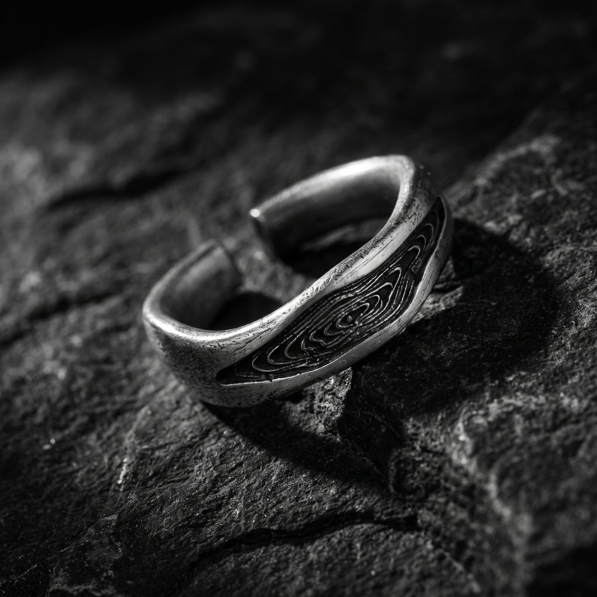 Angled view of the adjustable silver wood grain ring resting on dark rough stone.