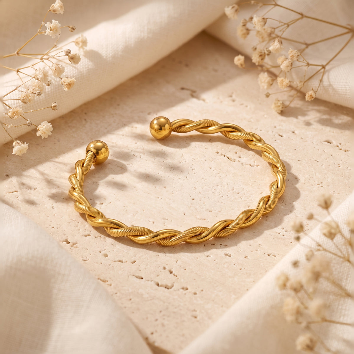 Angled perspective of the gold-tone twisted open cuff bracelet on a stone tile framed by soft fabric.