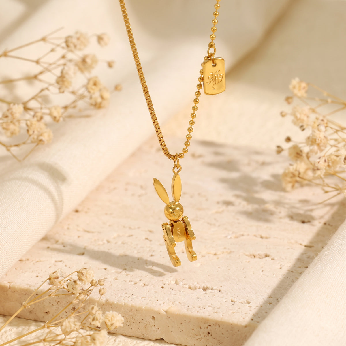 Gold articulated rabbit pendant necklace suspended above a textured stone slab with dried flowers.