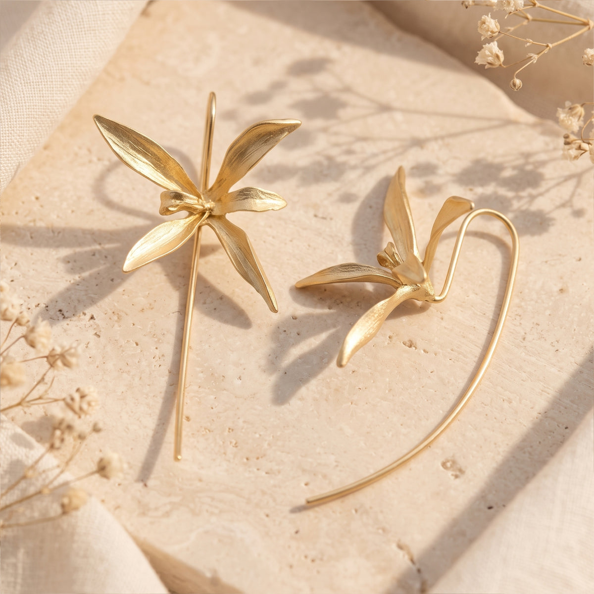Close-up of two gold-finish orchid drop earrings resting on a beige stone slab with soft shadows.