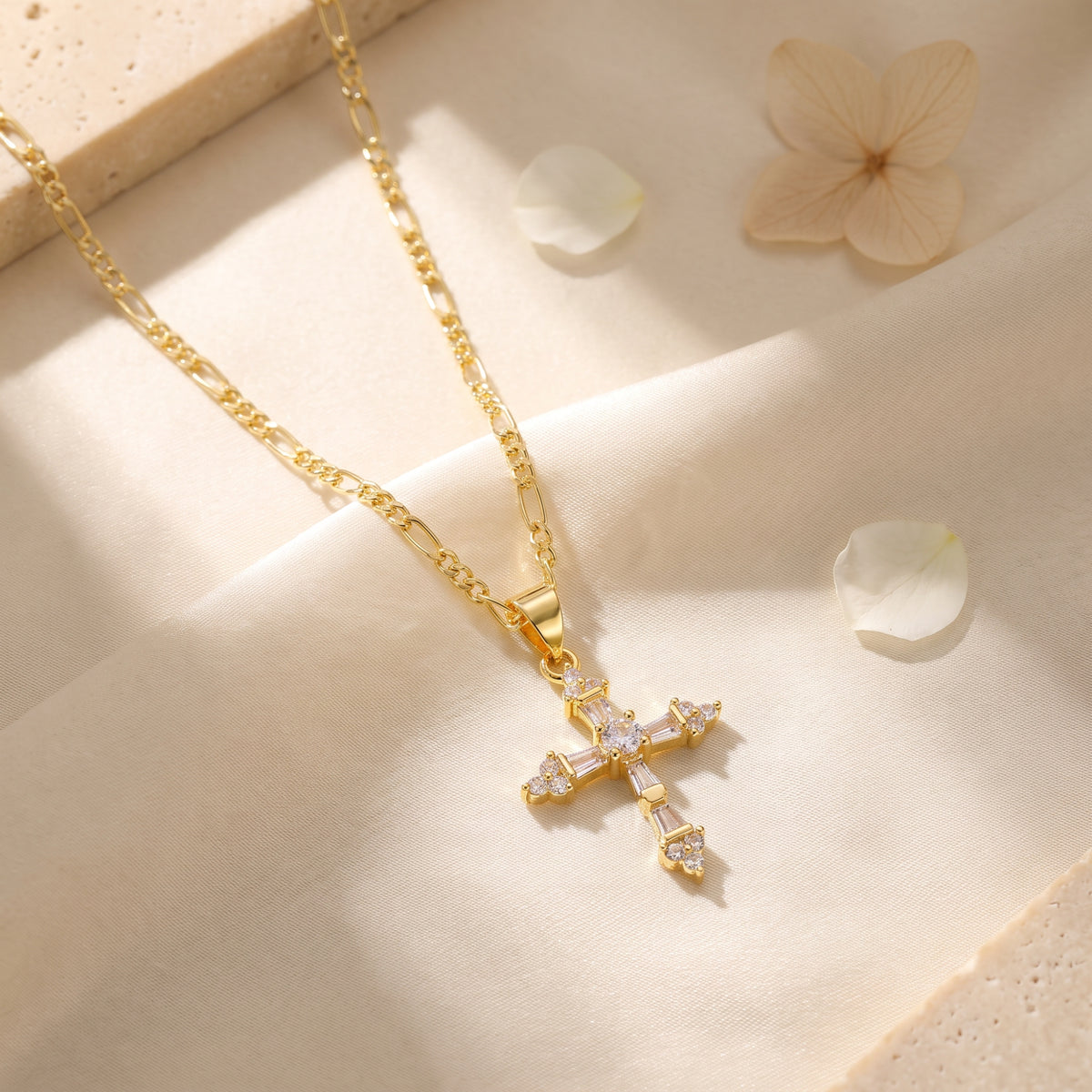 Gold-tone cross necklace resting on draped cream fabric with flower petals nearby.