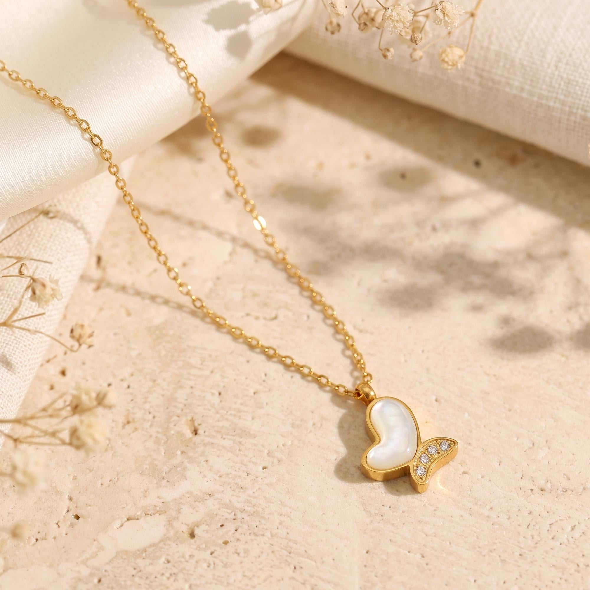 Gold butterfly necklace with mother of pearl inlay styled on a stone surface with dried flowers.
