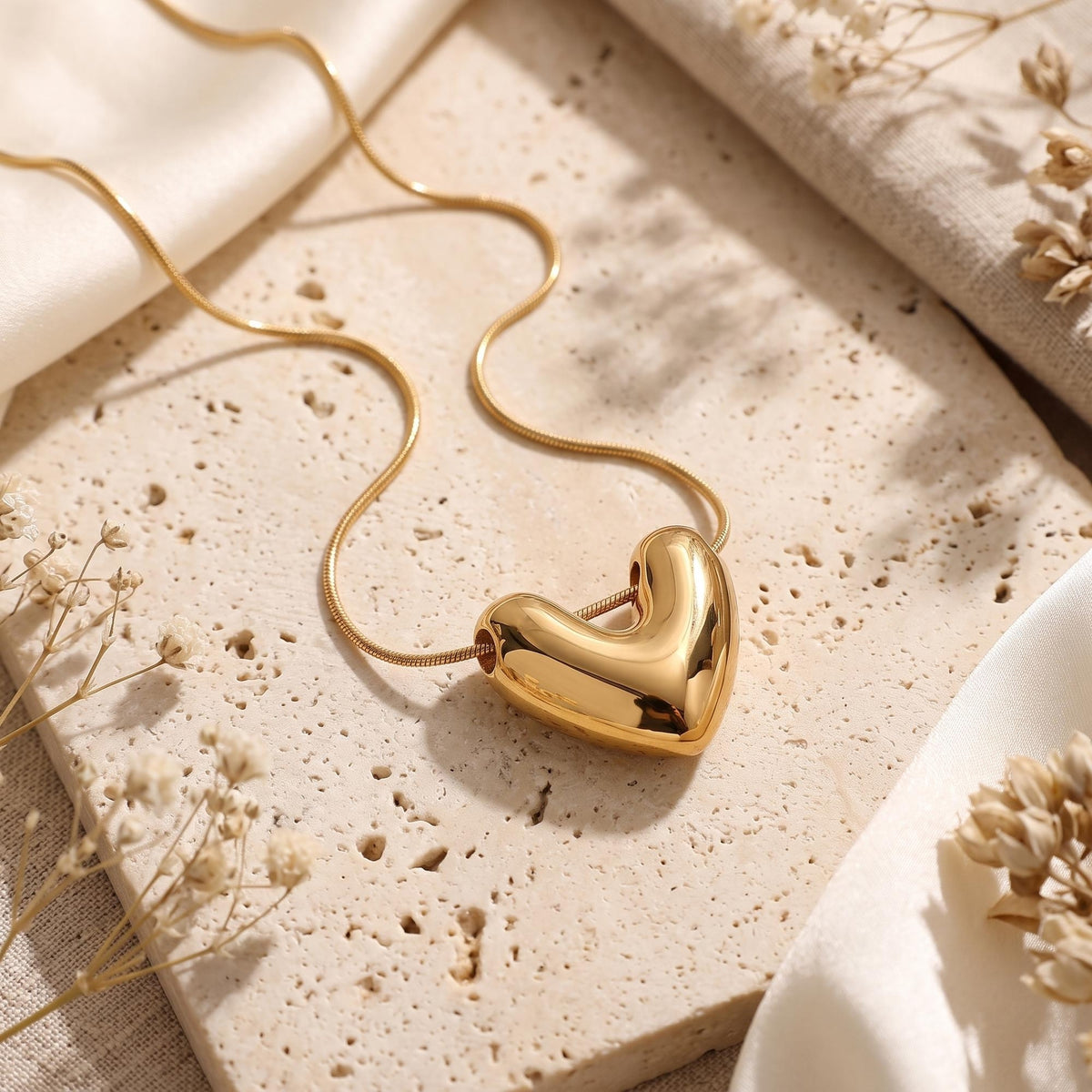 Gold-tone puffy heart pendant necklace arranged on a light travertine slab with dried flowers.