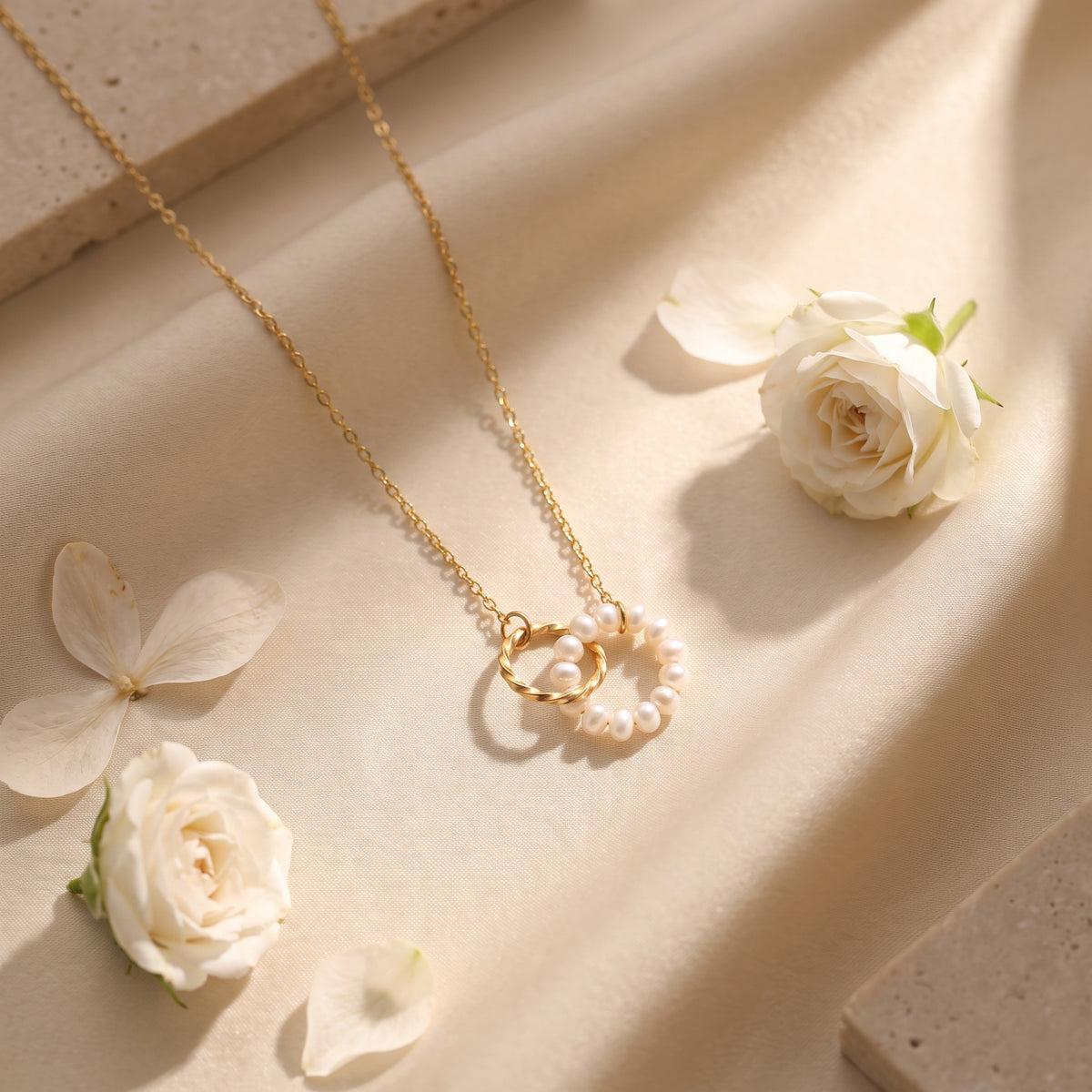 Flat lay of the gold-tone necklace with pearl circle and twisted ring pendant on cream fabric among white flowers.