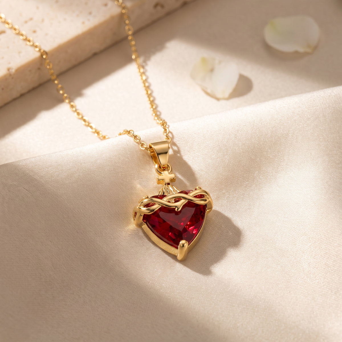 Close-up of the gold-tone pendant showing a faceted red heart crystal, intertwined crown detail, and cross on pale fabric.