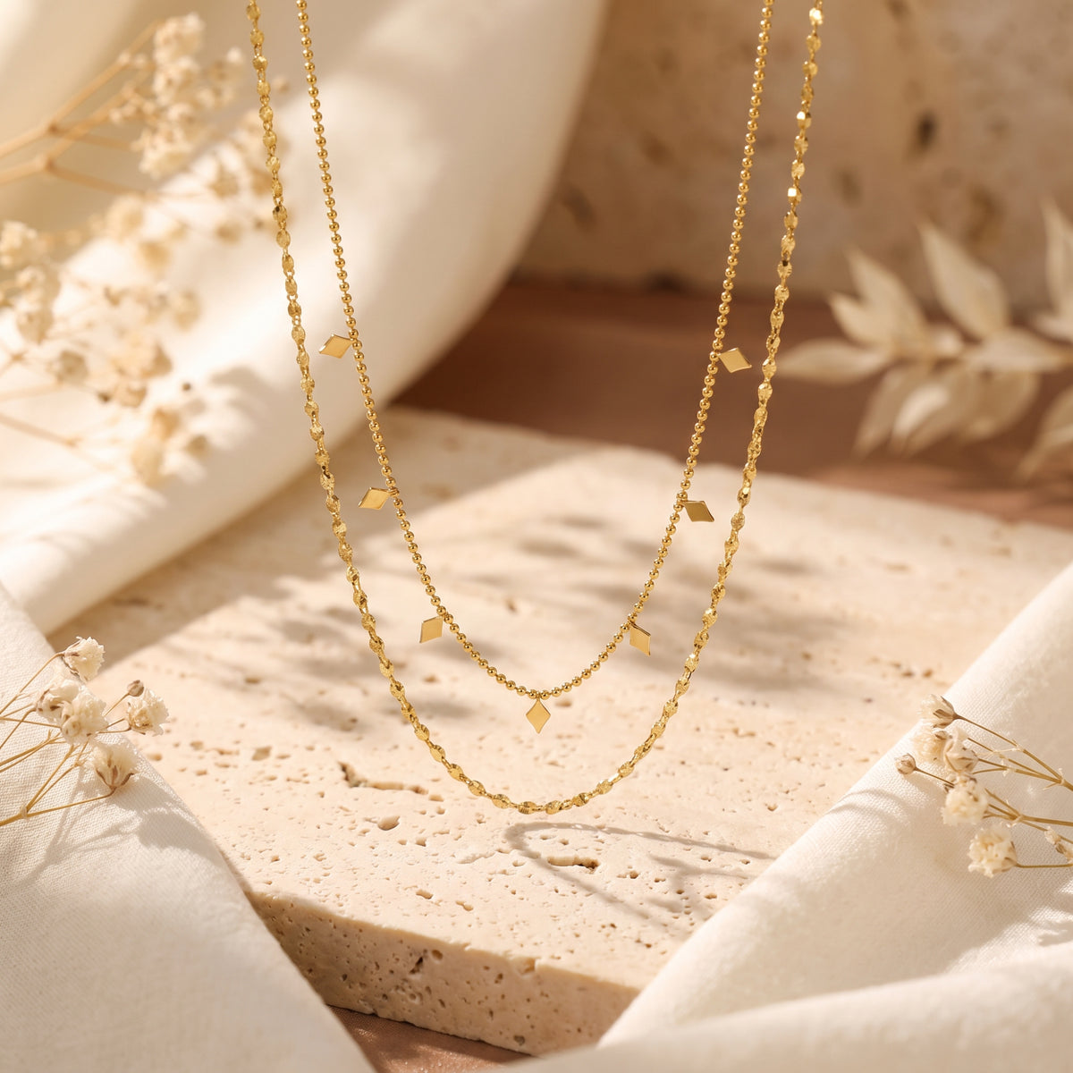 Gold-tone layered necklace hanging vertically above a beige stone block, with diamond-shaped charms on the inner chain.