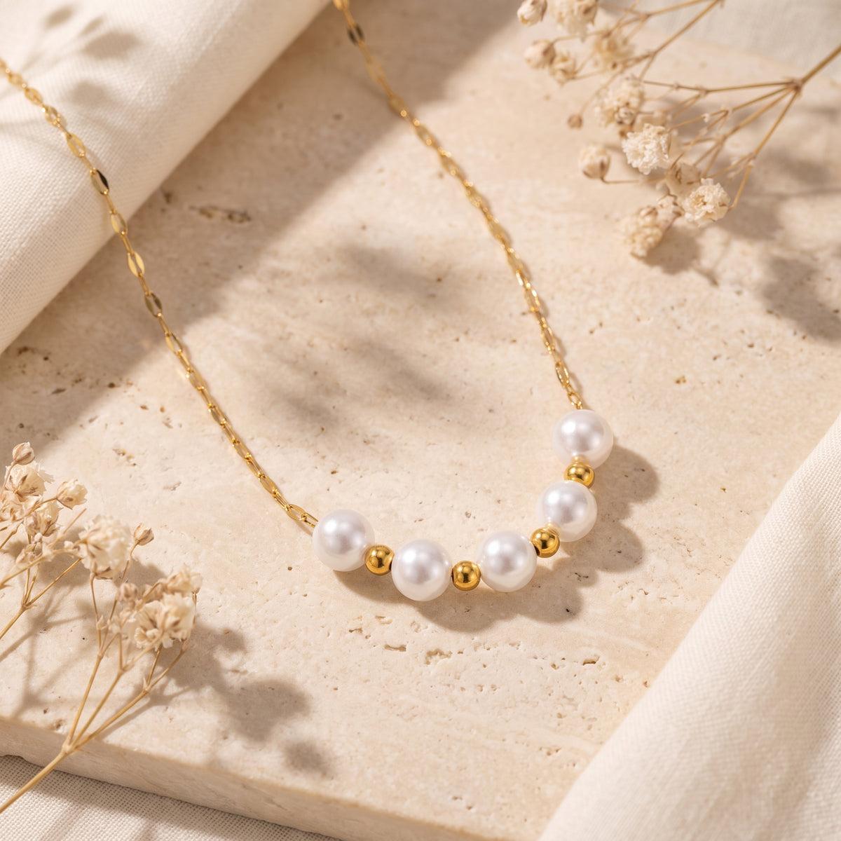 Flat lay view of the pearl station necklace resting on a textured beige stone block with delicate dried flowers.