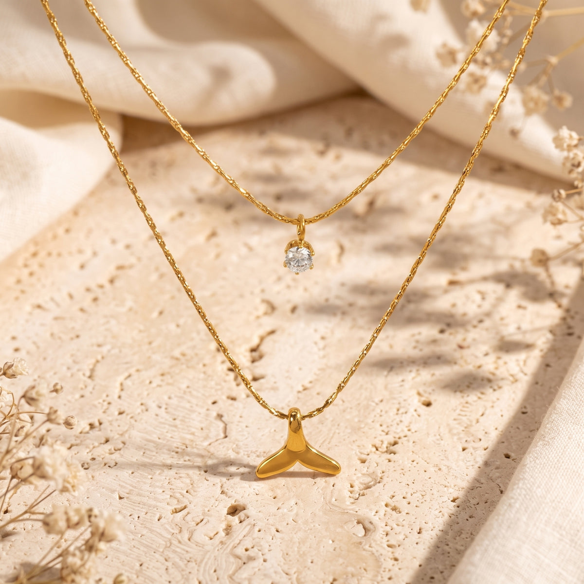 Gold-tone double-layered necklace arranged on a textured stone surface with crystal and whale tail pendants.