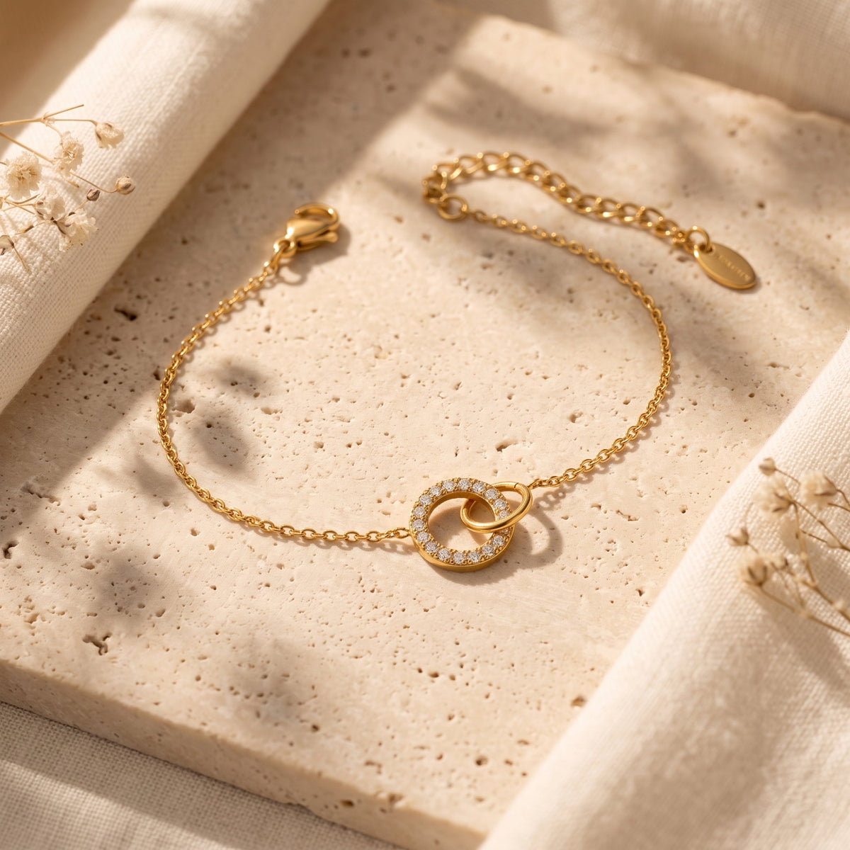 Gold-tone chain bracelet arranged diagonally on a textured beige stone slab, with interlocking circular centerpiece rings.