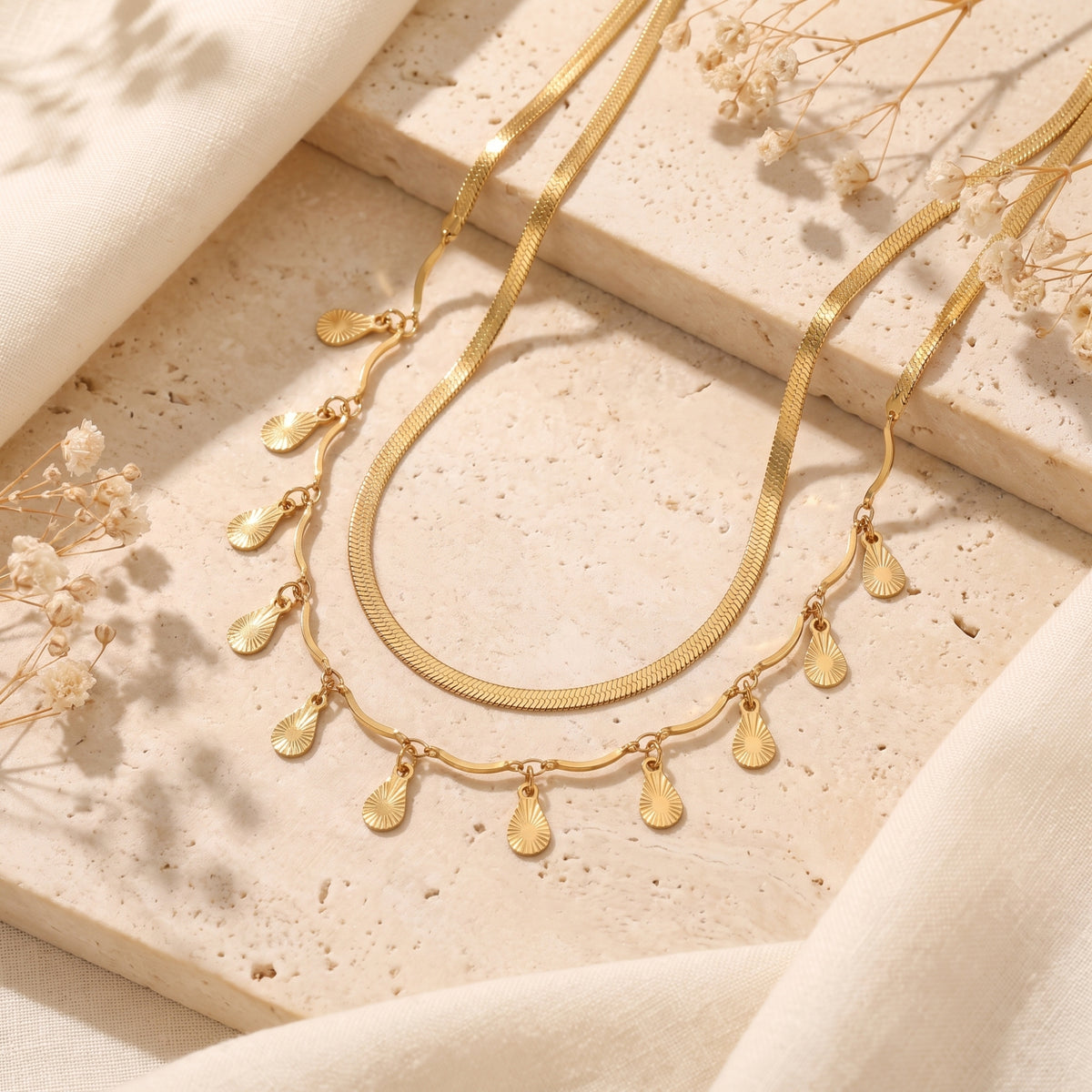 Gold-toned double-strand necklace with fan-pattern teardrop dangles resting on a light beige stone surface.