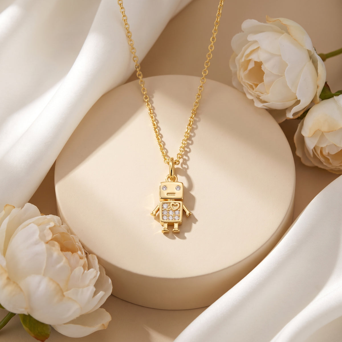 Gold-tone robot pendant necklace displayed on a round pedestal with white flowers and draped white fabric.