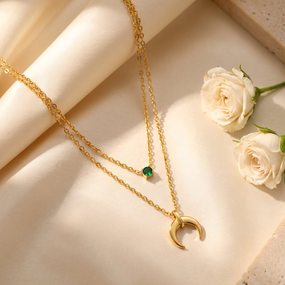 Flat lay of the gold-tone layered necklace with green cubic zirconia and crescent pendant on cream fabric beside white roses.