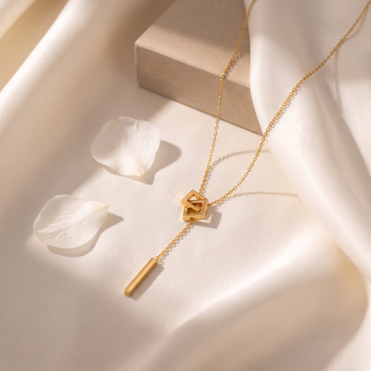 Gold-color square link lariat necklace laid flat on draped white silk fabric beside white petals.