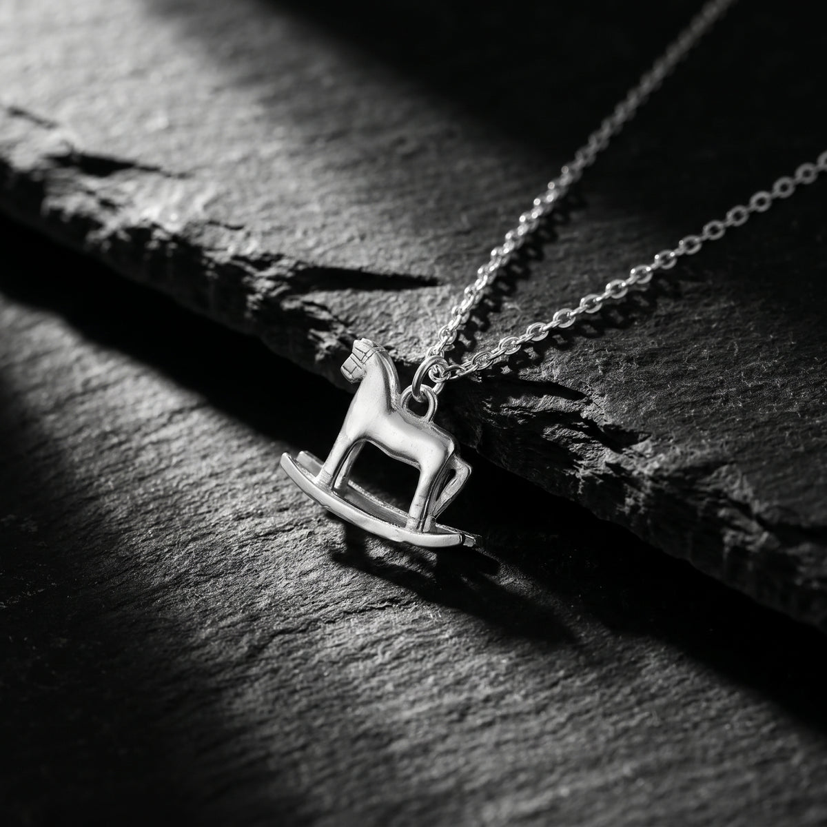 Angled close-up of the silver rocking horse pendant resting on a dark textured surface with the chain extending diagonally.