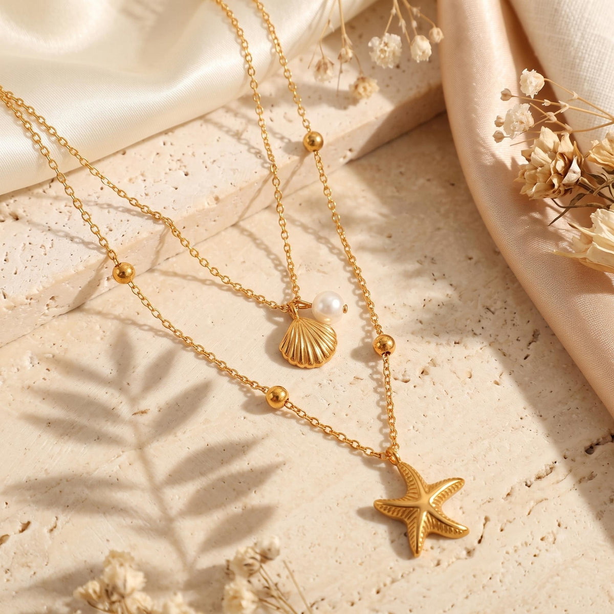 Flat lay of the gold-tone layered necklace with shell and starfish pendants on a beige stone surface with dried flowers.