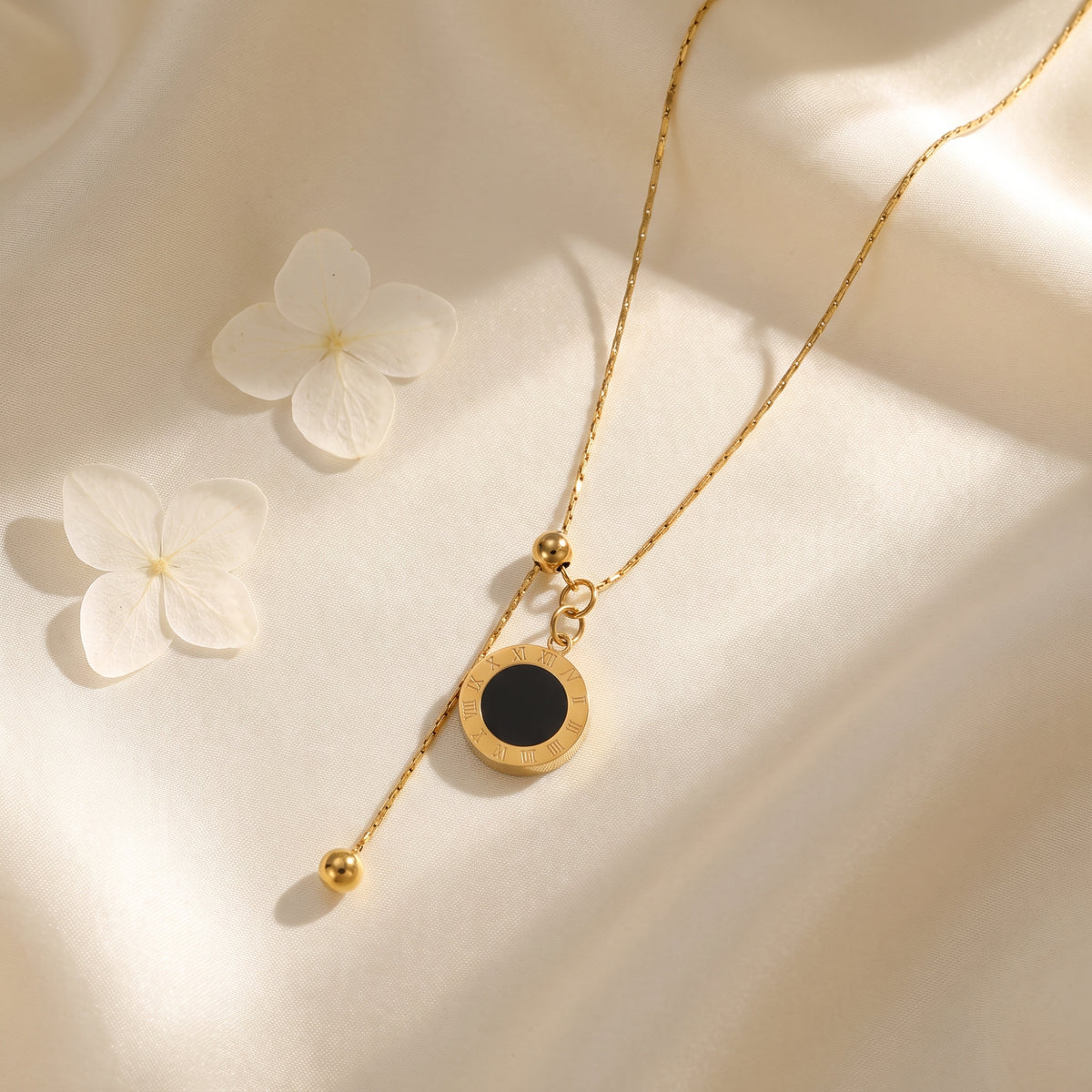 Gold-tone lariat necklace with black Roman numeral disc pendant arranged on white satin beside two white flowers.