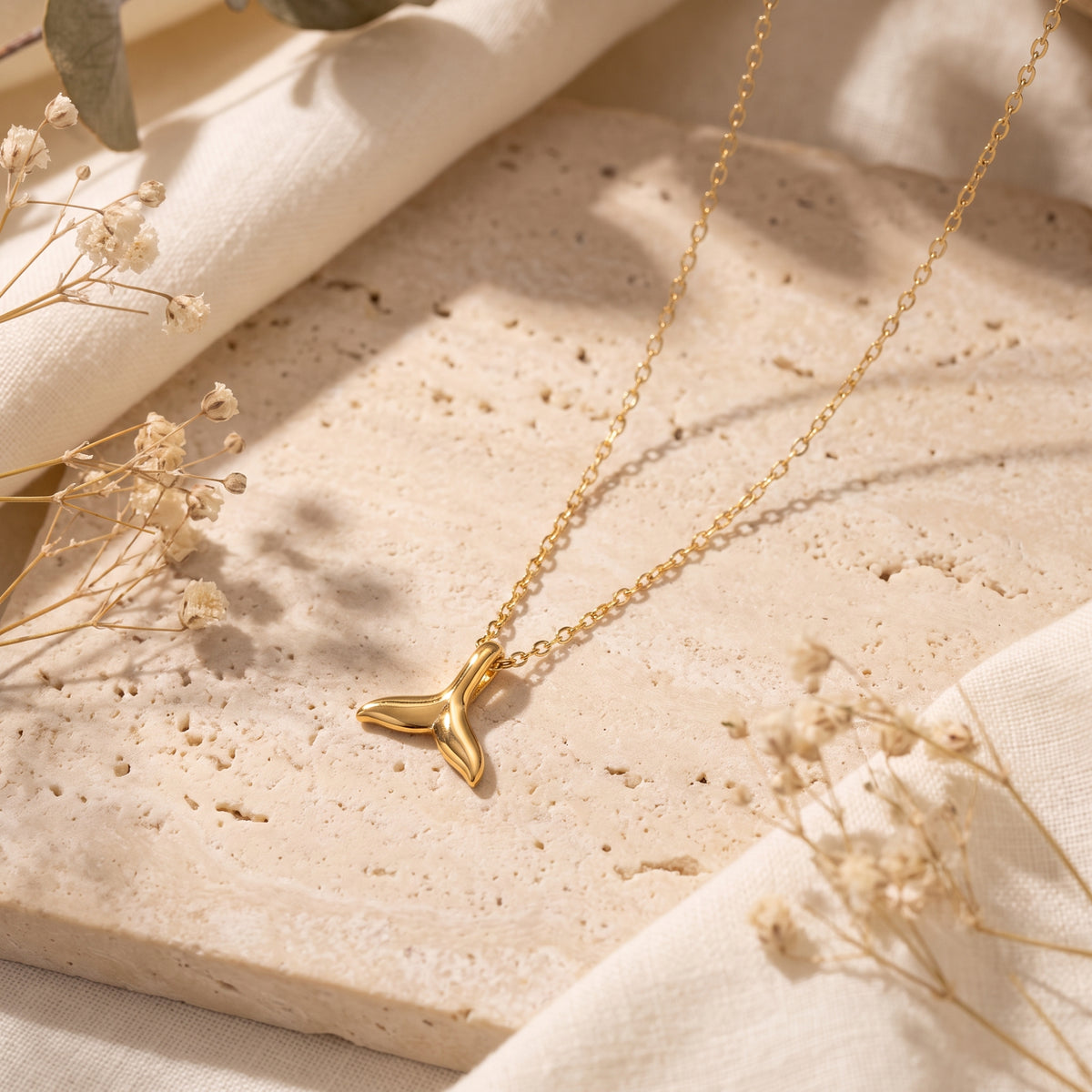 Gold-tone whale-tail pendant necklace lying diagonally on a beige stone slab beside dried flowers.