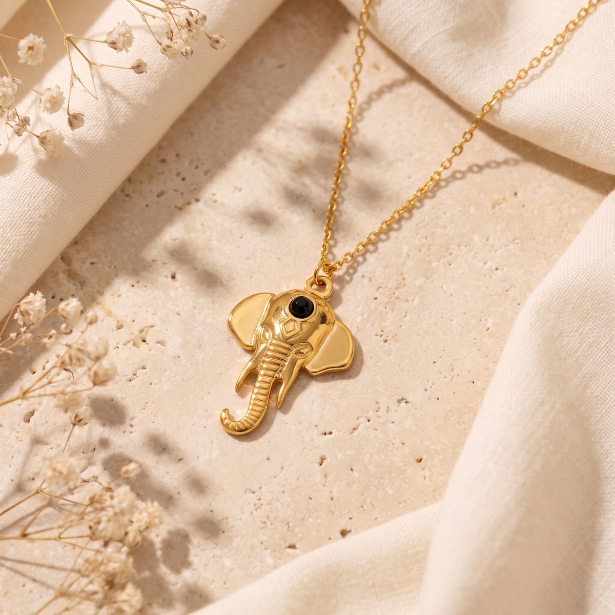 Flat lay of a gold-tone elephant pendant necklace on beige stone with light fabric and dried flowers.