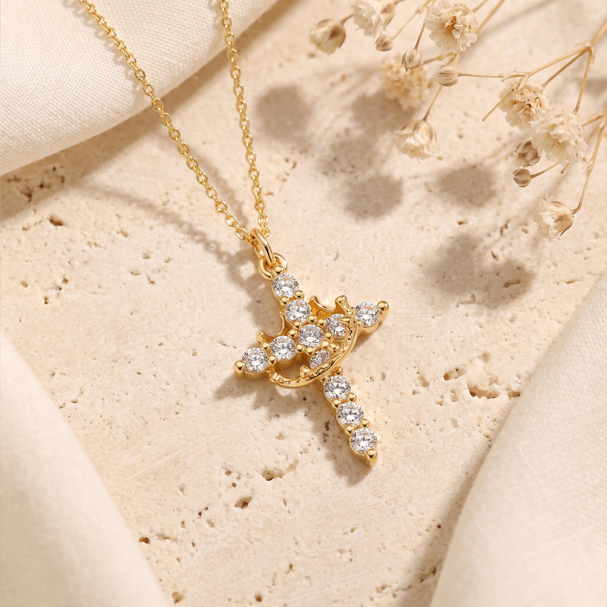 Angled laydown view of the gold-tone crown cross necklace on beige stone with dried flowers.