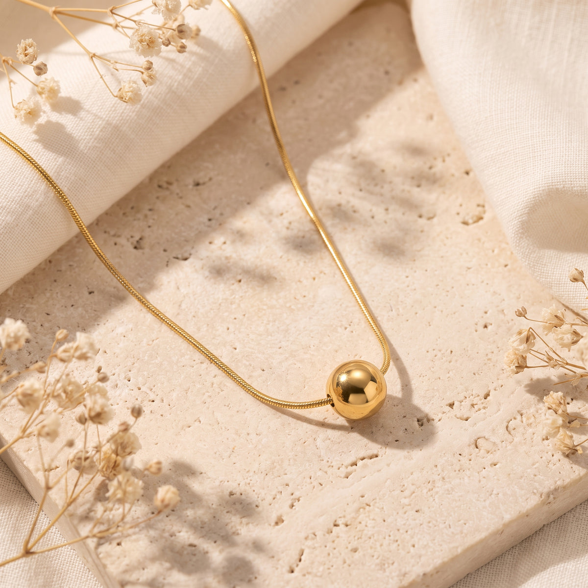 Flat-lay gold-tone sphere necklace on a beige stone slab with dried white flowers.