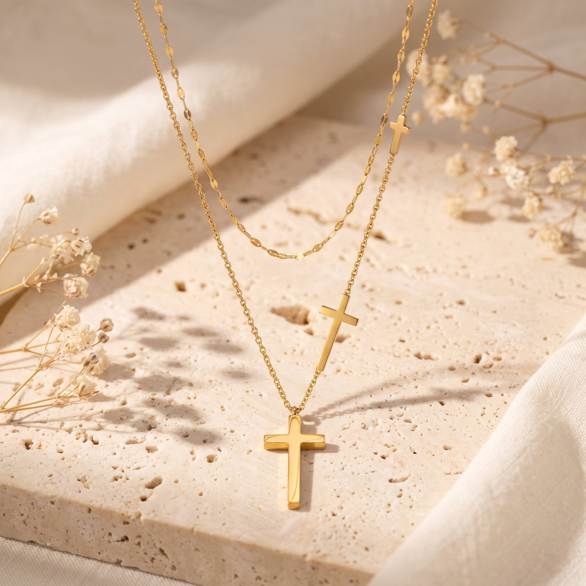 Gold-tone layered necklace with three cross pendants draped over a pale textured stone slab with dried flowers.