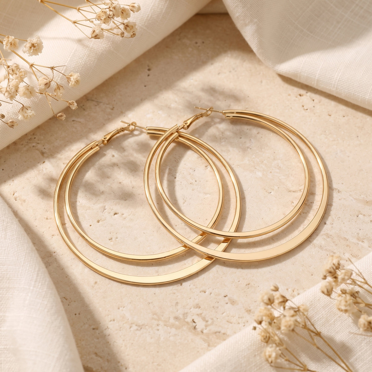 Pair of gold-tone double hoop earrings displayed flat on a textured beige stone surface with fabric and dried flowers.