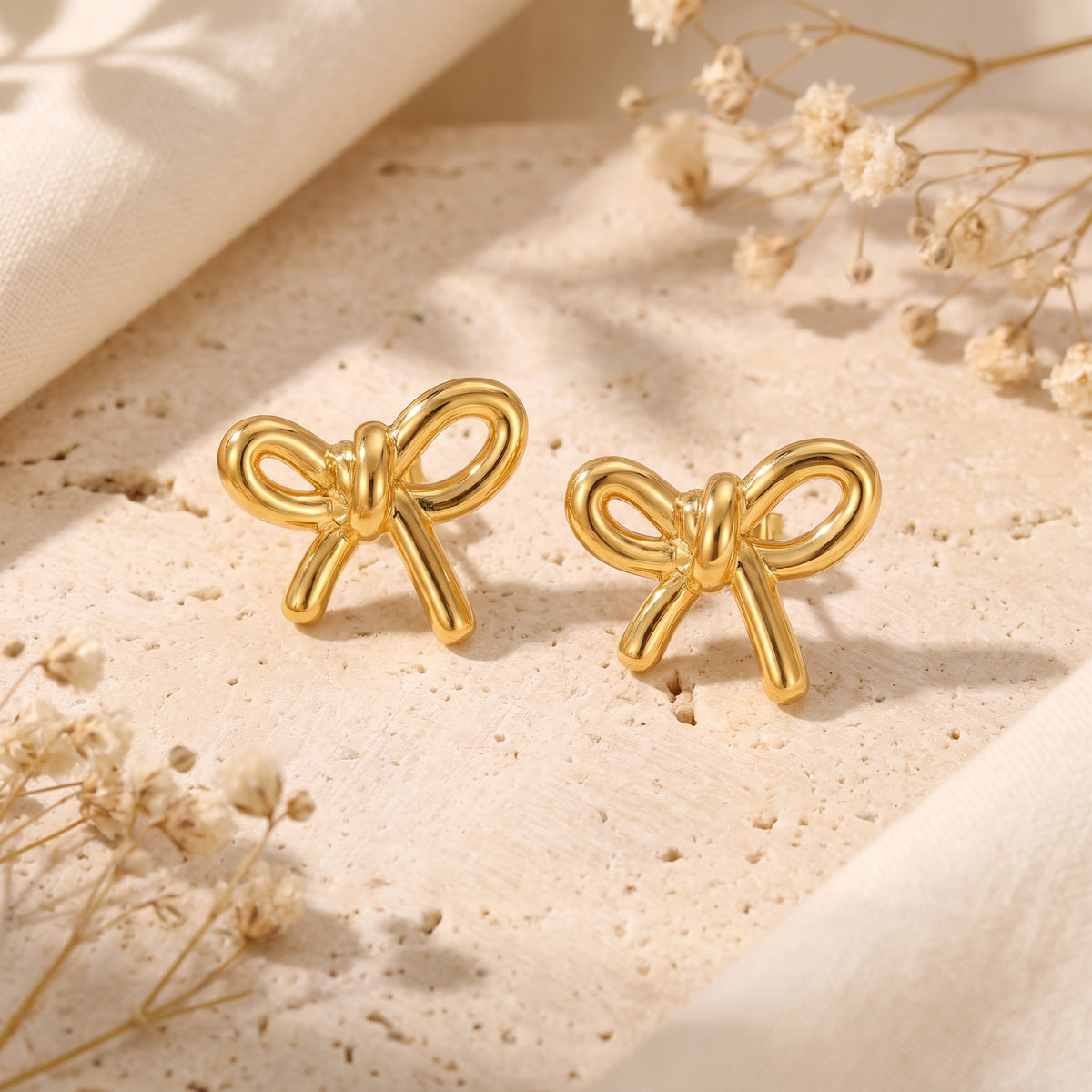 Close-up pair of gold-toned bow drop earrings displayed on a textured pale surface.