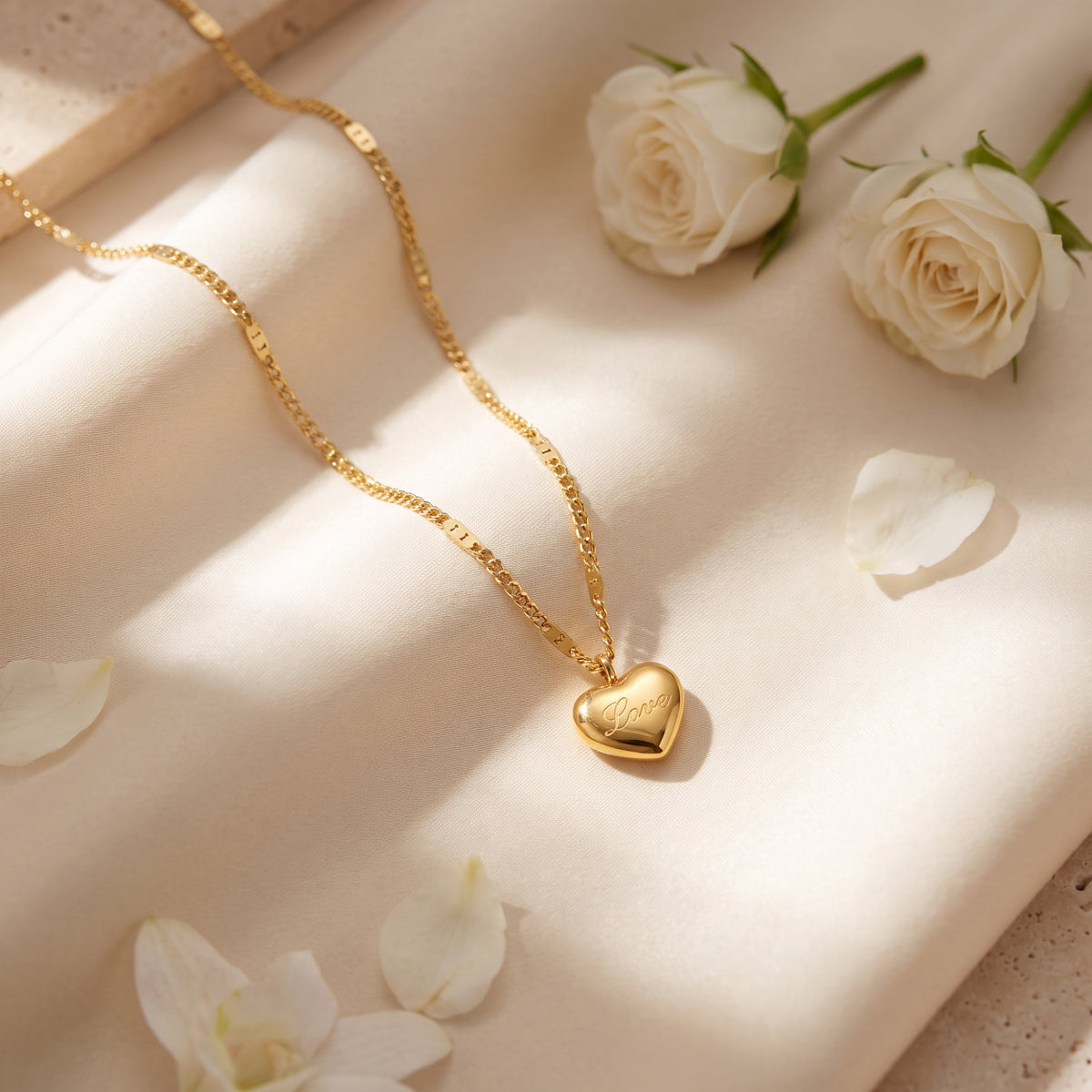 Gold-tone Love heart pendant necklace displayed on cream fabric with white roses and scattered petals.