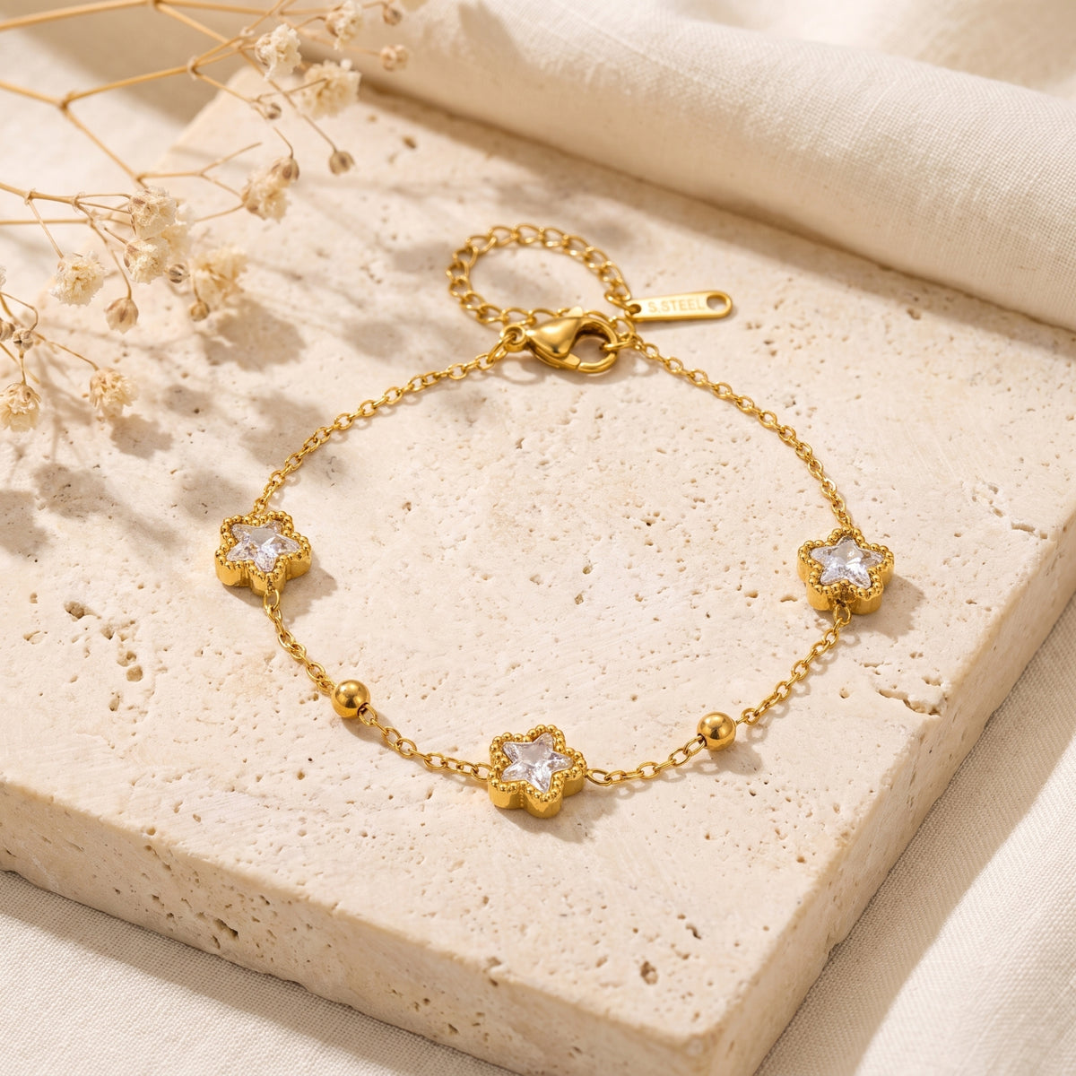 Flat-lay gold-tone station bracelet with three star-shaped cubic zirconia stations on a beige stone tile.